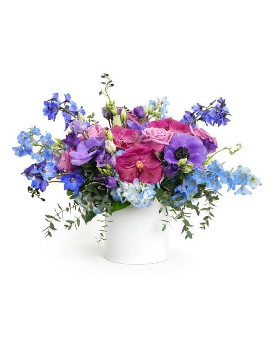 Vibrant floral arrangement featuring purple orchids, blue delphiniums, and lush greenery in a white vase.