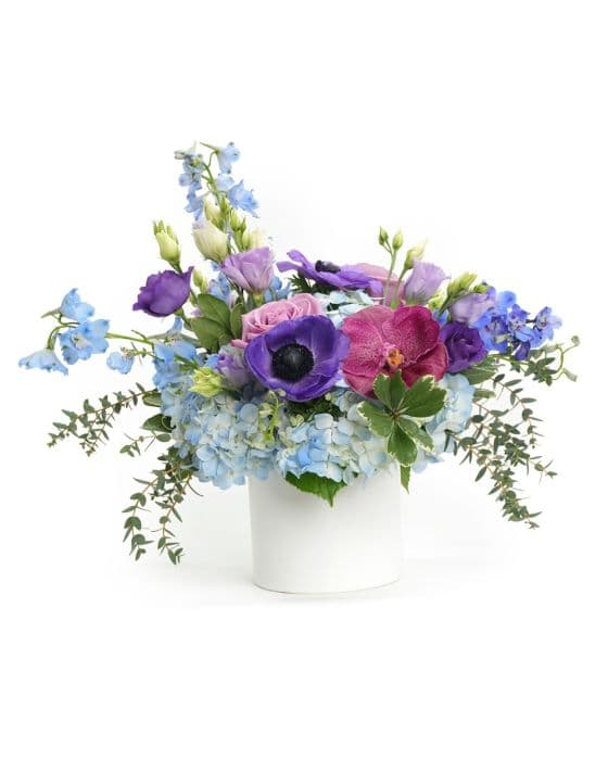 A vibrant floral arrangement featuring blue hydrangeas, purple orchids, and leafy accents in a white vase.