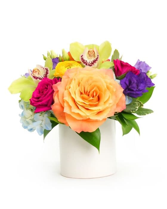 Vibrant mixed flower bouquet featuring coral roses, orchids, and seasonal blooms in a stylish white vase.