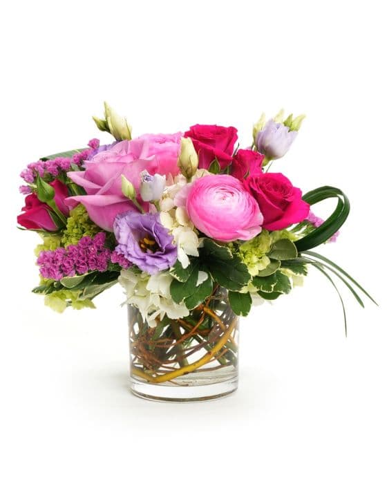 Vibrant floral arrangement featuring pink roses, purple tulips, and lush greenery in a clear glass vase.