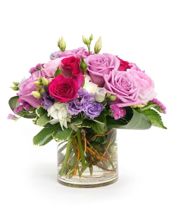 A vibrant bouquet featuring pink and purple roses, lush greenery, and delicate blooms in a clear glass vase.